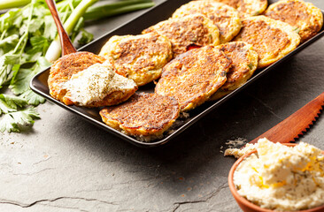 Turkish style zucchini pancakes or fritters known as mucver, served on black plate with fresh greens and sour cream. Close up image on dark stone background.
