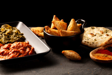 Close up image of a table with delicious dishes from India. Low light dark background with naan, curry chicken, palak paneer, samosa, various spices and samplers on stone