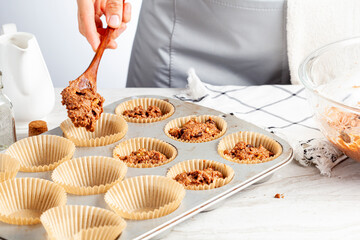 making of delicious fruit and nut cupcake on white marble countertop background. Muffin tin with liner, ingredients, and utensils are seen. A woman is adding mixture into the holes - obrazy, fototapety, plakaty