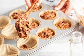 making of delicious fruit and nut cupcake on white marble countertop background. Muffin tin with liner, ingredients, and utensils are seen. A woman is adding mixture into the holes