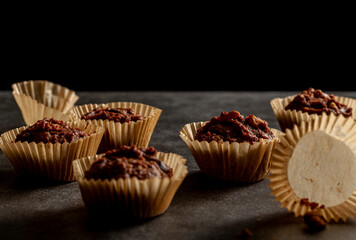 Shallow depth of field angled view of chocolate and nut muffins in liner papers fresh out of oven. Dark background moody low light image. Crumbles on stone background