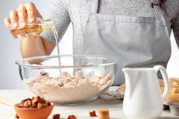 A woman is adding ingredients for making delicious fruit and nut cupcake on white marble countertop background. Muffin tin with liner, ingredients, and utensils are seen. Caucasian woman wearing apron