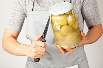 A caucasian woman wearing apron is trying to open a stubborn lid. She is inserting a sharp kitchen knife between the jar and the lid to relieve pressure and unscrew the tight lid.