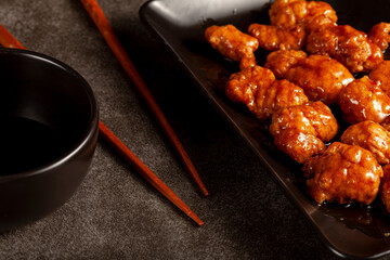 mandarin orange chicken served on black porcelain plates. Special savory sauce is added using spoon . Chopsticks are seen nearby. A Chinese dish popular as a snack in America.