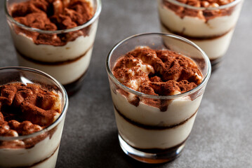 Delicious traditional tiramisu desert served in small glass cups on stone countertop. Low light image shows texture and layers from angle with shallow depth of field.