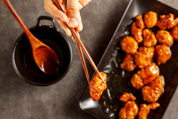 A person is eating delicious mandarin orange chicken, a Chinese American dish using wooden chopsticks. The sauce is served in porcelain bowl. Dark background. Top view close up image