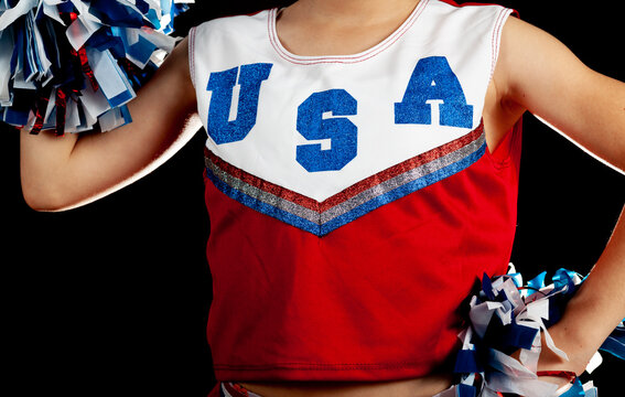 A caucasian teen girl wearing sleeveless shirt with USA printed on top against dark background is holding two pom poms. One hand on waist the other above Cheerleading for the national team concept.
