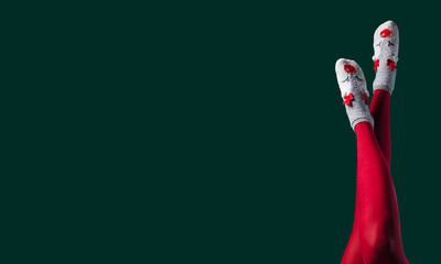 Christmas banner with women legs in red tights and funny warm socks with reindeers. Female feet on dark green background with copy space for text.