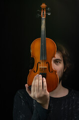 Fototapeta premium The eye of a young dark-haired man peeking out from behind an old violin. Dark background. Selective focus.