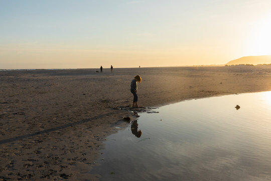 Boy on a beach, looking at his reflection in a water pool, low tide, sunset.