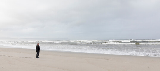 A man walking across sand to the water's edge on a beach, overcast day and surf waves breaking on shore. 