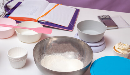 Layout with digital scales, notepad, bowls on a white table. In the foreground is a bowl of flour and a cupcake tin. Selective focus.