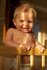 An incredibly beautiful baby is standing in a crib and smiling happily at sunset