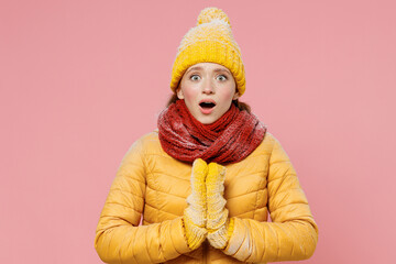 Shocked surprised young woman 20s years old wears yellow jacket hat mittens holds hands folded in prayer gesture begging about something isolated on plain pastel light pink background studio portrait.