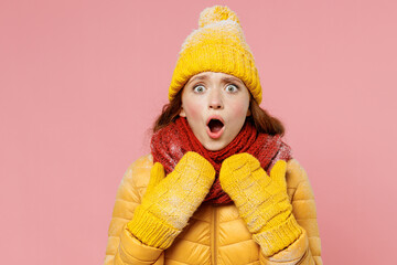 Frightened afraid scared amazed young woman 20s wears yellow jacket hat mittens look camera keeping mouth wide open point hands herself isolated on plain pastel light pink background studio portrait.