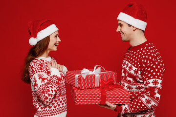 Young side view fun couple friends two man woman wear sweater hat give red present box with gift ribbon bow isolated on plain red background. Happy New Year 2022 celebration merry ho x-mas concept