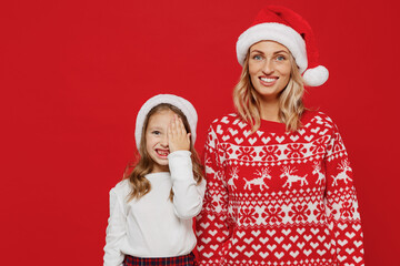 Young woman wearing sweater hat have fun with child baby girl 6-7 years old. Mommy little kid daughter cover eye with hand isolated on plain red color background studio. New Year love family concept.
