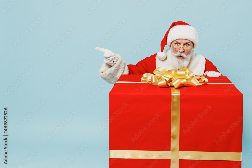 Wall mural Full size old Santa Claus man in Christmas hat red suit clothes stand near big gift box point finger aside isolated on plain blue background studio. Happy New Year 2022 merry ho x-mas holiday concept