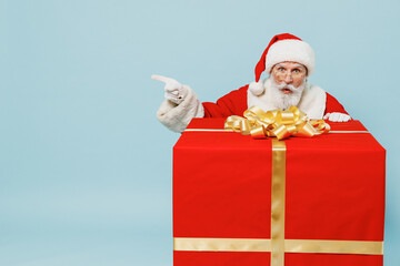 Full size old Santa Claus man in Christmas hat red suit clothes stand near big gift box point finger aside isolated on plain blue background studio. Happy New Year 2022 merry ho x-mas holiday concept