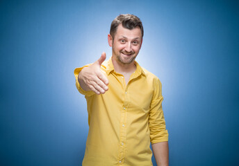 Smiling young man giving hand for handshake over blue background, dresses in yellow shirt. Greeting gesture concept