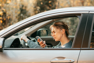 Naklejka premium Young beautiful woman drives car, violates traffic rules, gets distracted, looks at screen of mobile phone, uses smartphone, fast paced modern life concept