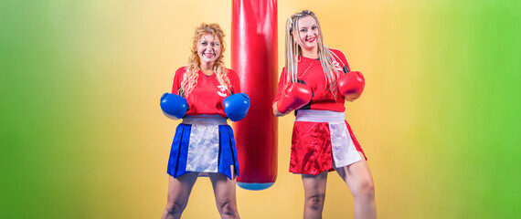 Two young girls, mixed martial arts fighters, in bright tracksuits and boxing gloves, stand against punching bag and yellow background. They look into camera and smile.