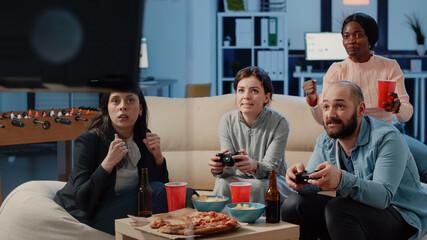 Coworkers playing video games with joysticks and losing after work at office. Colleagues enjoying game with controllers on television console for entertainment and drinks. People having fun