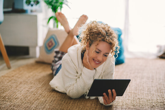 Happy adult woman laying on the carpet floor at home using modern tablet and internet connection wireless at home. Female people enjoy indoor relax leisure activity alone watching web device