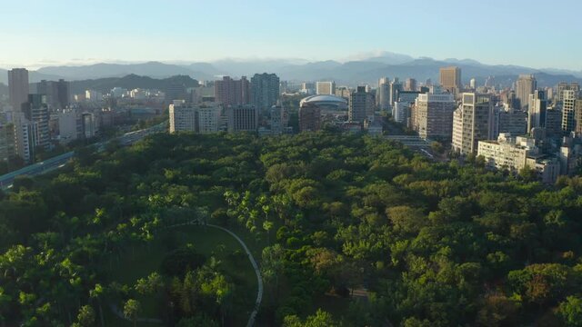 Epic Aerial flyover rural Daan Park with green trees surrounded by skyline with skyscraper buildings and silhouette of mountains in background - Taipei City,Taiwan during daytime and sun