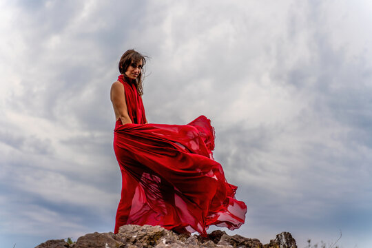 A woman in a red dress stands above a stormy sky, her dress fluttering, the fabric flying in the wind.