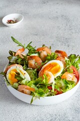 Fresh crispy salad with vegetables, with seafood (shrimps) and poached egg in a white plate on a light background. Healthy food concept.