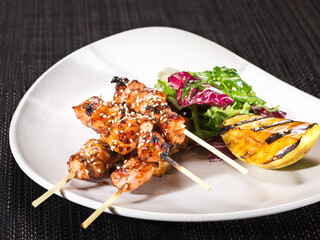 portion of grilled chicken fillet skewers with mix salad and grilled lemon slice on a plate on dark background