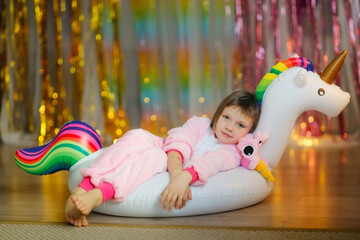 Cute kid in a pink soft unicorn costume plays with an inflatable unicorn ring on a colorful bright background, pajama theme party