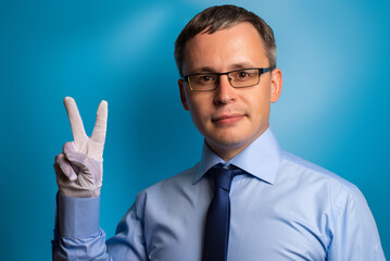Smiling man in blue shirt and tie shows the V sign with his hand in a white glove. Confident specialist on a blue background.