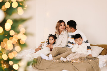 A mixed race Asian family with two children in cozy sweaters is relaxing together, resting and having fun on a bed in a decorated bedroom with Christmas tree at home in the New Year. Selective focus