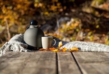 there is a thermos and a mug of hot tea with a gray blanket and yellow leaves on a wooden table in autumn in nature
