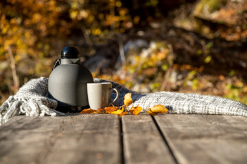 there is a thermos and a mug of hot tea with a gray blanket and yellow leaves on a wooden table in autumn in nature