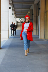 An attractive girl in a red beret and a red coat walking on the street in a  city. Woman in jeans, brown boots. Fashion portrait, casual model goes, looks straight and smiles. Blurred details