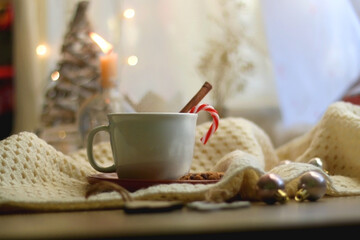 Cup of warm drink with candy cane and cinnamon stick, soft blanket and various Christmas decorations. Hygge at home. Selective focus.
