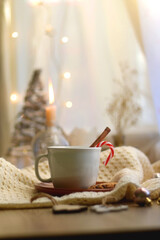 Cup of warm drink with candy cane and cinnamon stick, soft blanket and various Christmas decorations. Hygge at home. Selective focus.