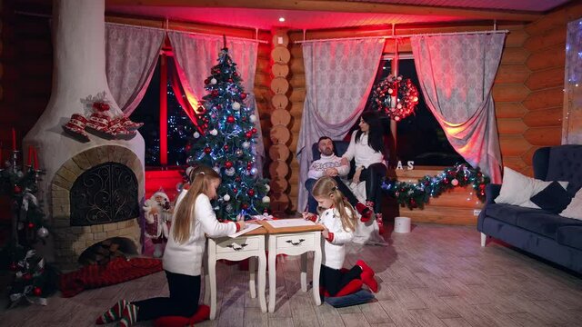 Beautiful family of four members having rest in a room ready for Christmas. Daddy and mommy are peacefully chatting on the armchair. Girls are drawing pictures at the fir-tree.