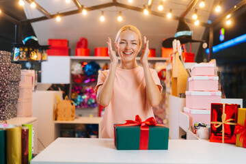 Excited happy young woman standing by counter table with wrapped Christmas gift box tied red ribbon and decorated beautiful bow looking at camera, bright blurred background of festive packages.