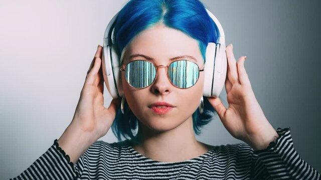 Electronic music. Digital people. Futuristic technology. Funky girl with blue hair in glitch flicker motion glasses listening mp3 sound in wireless headphones isolated on light.