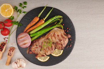 Grilled pork steak from a summer BBQ served with Vegetables, asparagus, baby carrots, fresh tomatoes and  spices. Grilled steak on  black slate on stone background . top view.