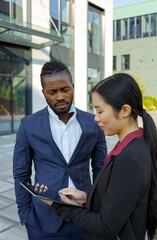 Colleagues African American man with stubble in formal suit and Asian young woman with long brunette hair check work using tablet PC