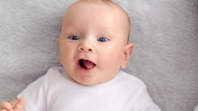 Child's face of a Happy newborn baby looking to camera.  Healthy newborn baby in a white t-shirt with blue eyes. Slow motion.  Face of a Beautiful active  tiny child. Cute white Infant boy, top view.