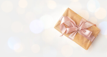 Obraz na płótnie Canvas Banner of birthday or New Year's eve present in kraft paper with pink ribbon on soft white background with bokeh lights. Xmas composition. Flat lay. Happy holidays celebration and giving love concept