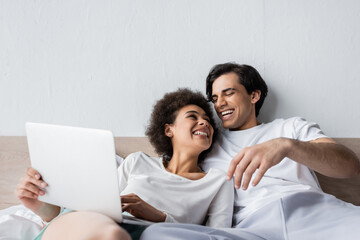happy african american freelancer using laptop near cheerful boyfriend in bed
