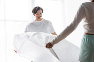 happy man arranging bed sheet with african american girlfriend