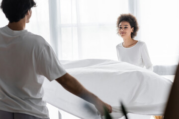 positive african american woman arranging bed sheet with boyfriend